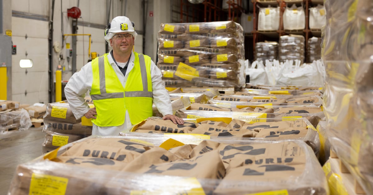 PURIS Dawson plant worker in yellow vest and white hat stands by pallets of packaged pea protein.